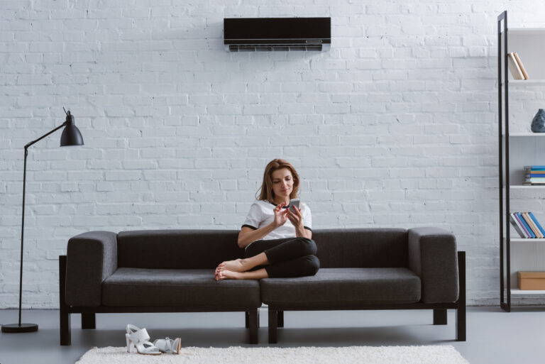 relaxed young woman using smartphone on sofa under air conditioner hanging on wall